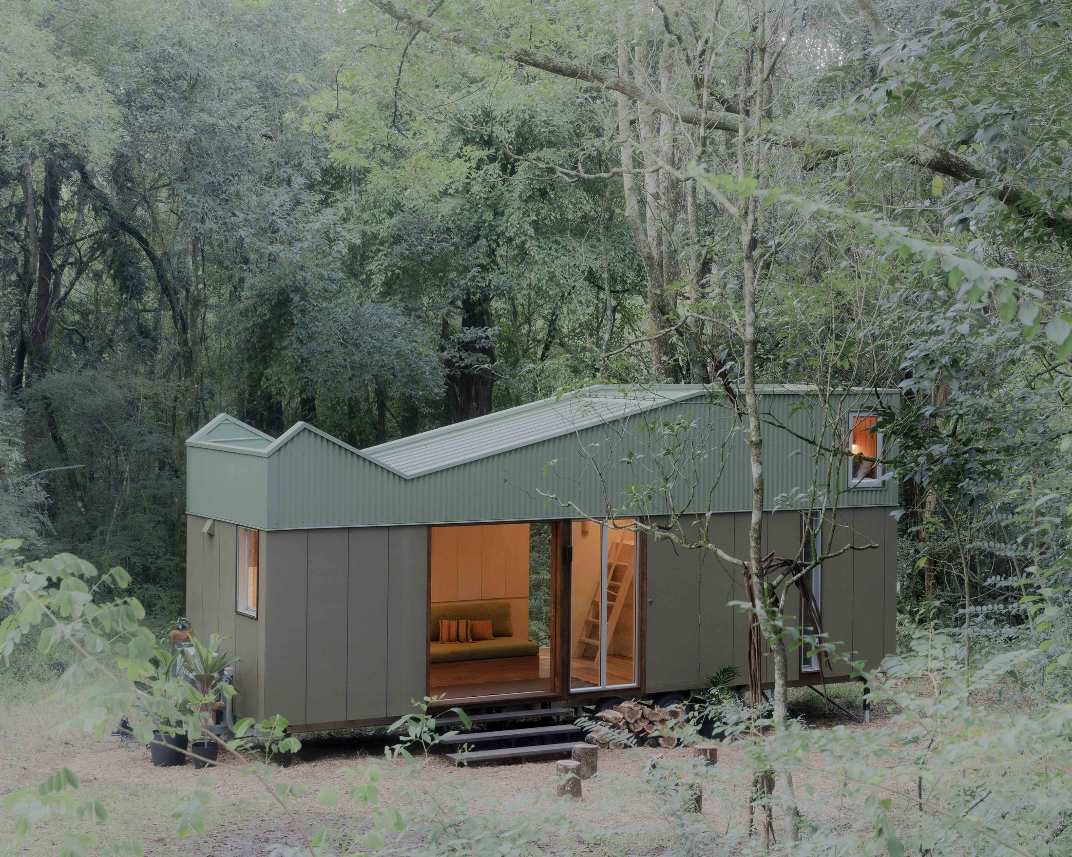 Rowan, a timber and steel tiny house nestled in a forest clearing at dusk, warm light glowing from inside