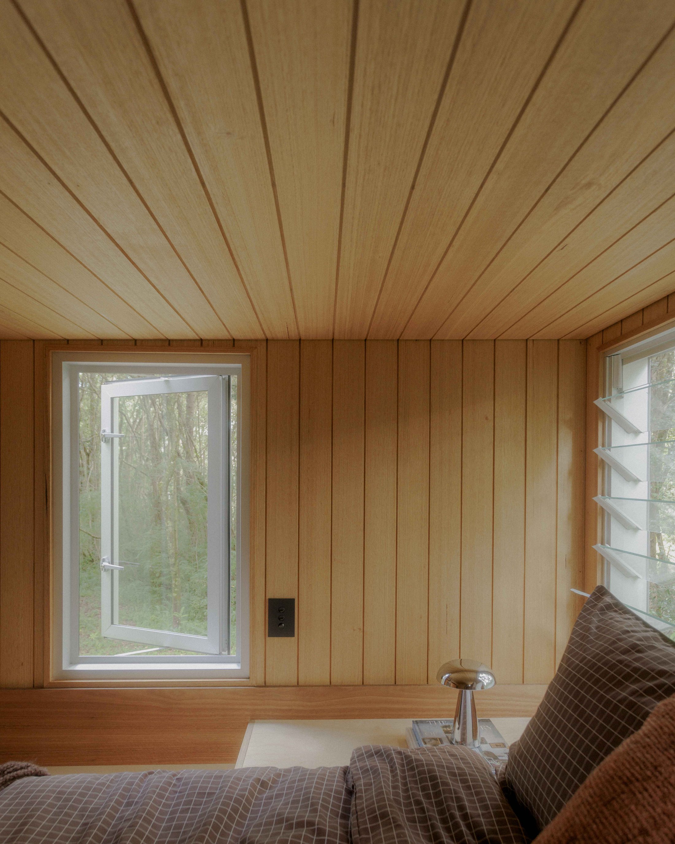 Open loft window looking out to forest, with bed and pillows in foreground
